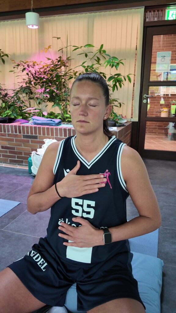 A woman in a black sports shirt sits inside, her eyes closed, her hand placed on her chest and stomach; she seems calm and focused – she may be practicing mindful breathing or meditation, as the thermal baths optimize your workout.