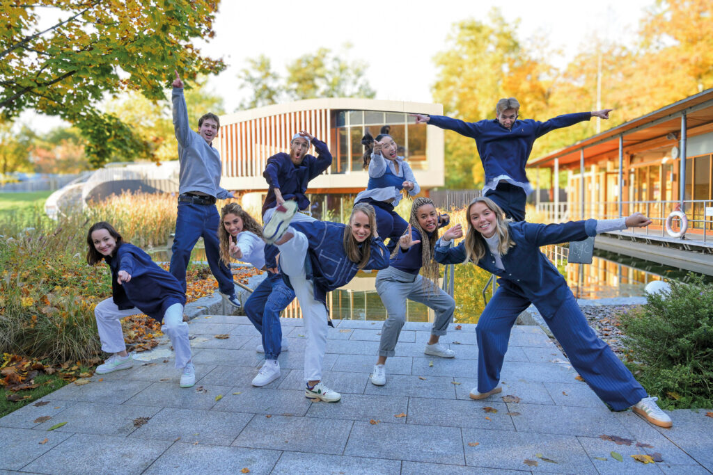 A group of nine people dressed in casual blue and gray clothing playfully pose outdoors on a paved path under the bright sun, with trees, grass, modern buildings, and a hint of Keidel in the full moon energy in the background.