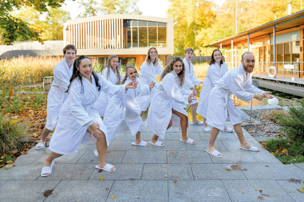 Nine people in white bathrobes and slippers playfully pose outdoors on a stone path, in the background modern buildings and autumn-coloured trees can be seen - smiling and laughing, they embody how "the thermal bath" optimises your training through relaxation and shared energy.