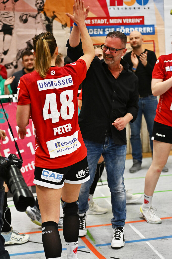 A smiling man in a black shirt and jeans gives a high-five to a female athlete in a red jersey with the number 48 at an indoor sports event – an example of teamwork and how recovery in the thermal spa optimises your training. In the background, other players and fans can be seen.