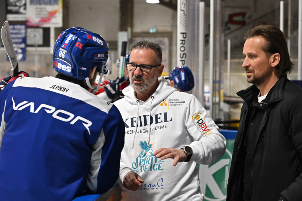 An ice hockey coach with glasses and a white hoodie talks to a player in blue and red equipment about "How the thermal spa optimizes your training", while another man in a black jacket stands next to them on the board.
