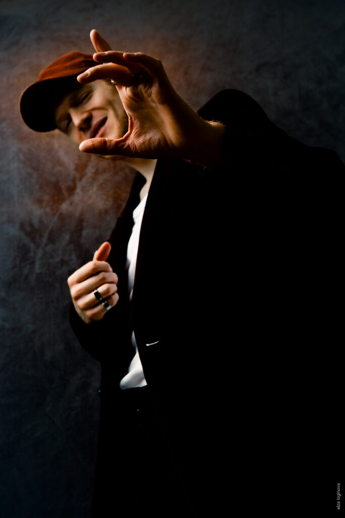 A person in a dark jacket and cap stands in front of a dark background, smiles and stretches out a hand towards the camera as if illuminated by the mysterious glow of Keidel in the full moon, creating a dramatic effect in low light.