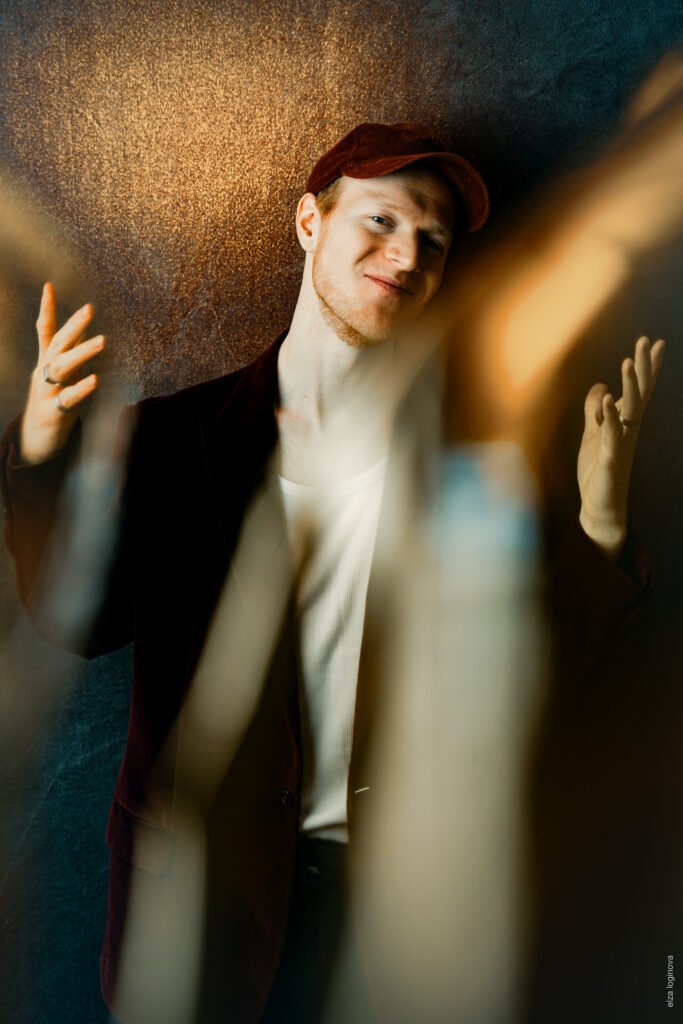 A man in a dark jacket, white shirt and red cap stands in front of a textured background and gestures with both hands, while warm streaks of light blur the foreground - an artistic effect reminiscent of Keidel in the full moon.