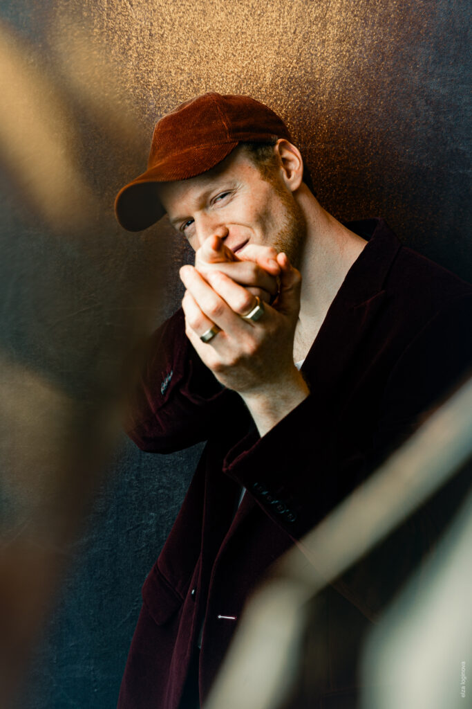 A man in a brown cap and dark blazer smiles subtly, holds his hands together in front of his face, and stands in front of a textured brown background, while the soft light highlights his features - a reference to the calm charm of Keidel in the full moon.