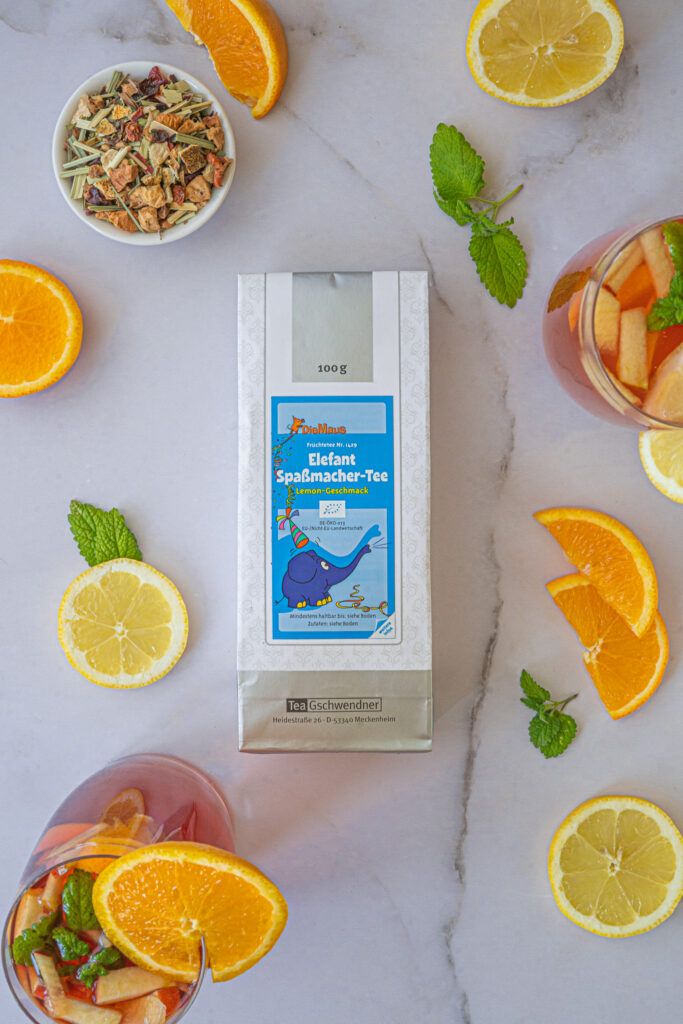 A tea pack stands in the middle of a bright surface, surrounded by orange slices, mint leaves, a loose tea blend inspired by children's saunas in a bowl and two glasses of fruit tea garnished with orange slices.