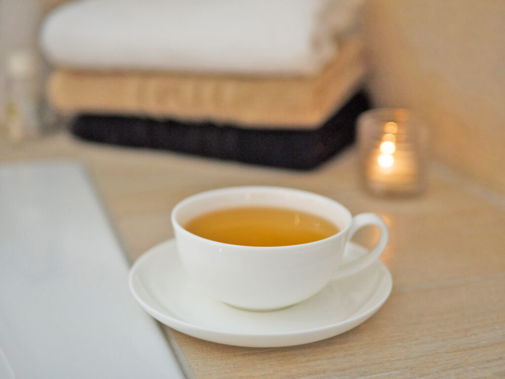 A white cup filled with tea stands with a matching saucer on a light wooden surface. In the background, folded towels and a small lit candle create a calm, relaxing atmosphere - perfect for enjoying Keidel in the full moon.