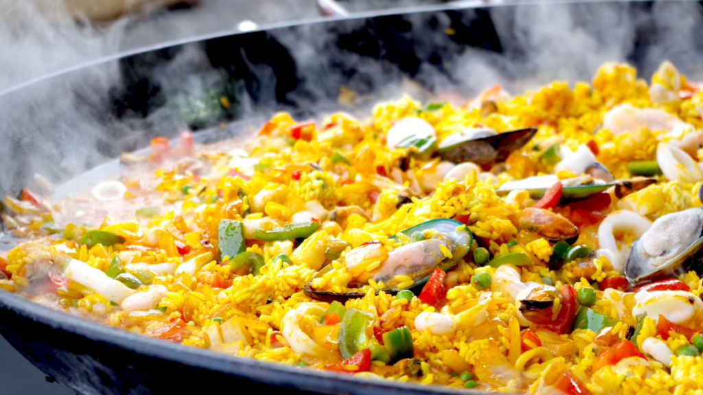 Close-up of a large pan of steaming seafood paella under the wedge in the full moon, with yellow rice, mussels, clams, shrimp, squid, peas and chopped red and green peppers.