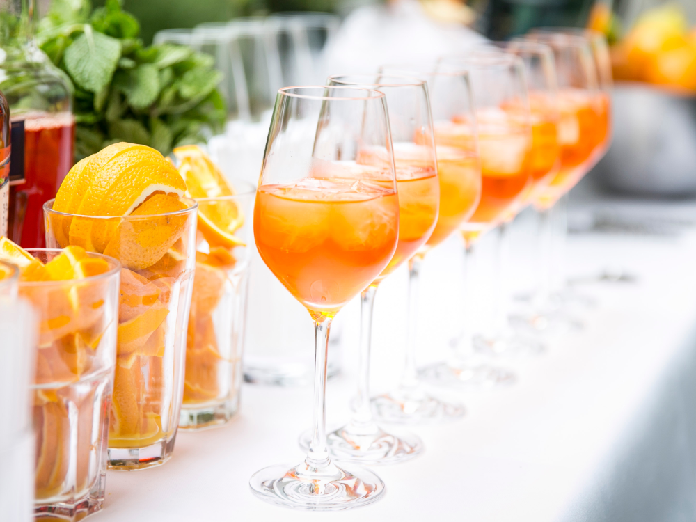 On a table are several wine glasses filled with orange cocktails and ice, as well as glasses with orange slices and fresh mint - perfect for enjoying the summer prepaid card on a sunny afternoon.