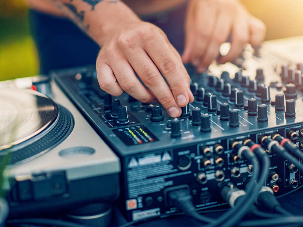 One person adjusts the buttons of a professional DJ mixer, one hand on the controls, the other near a turntable. The outdoor area in bright daylight captures the perfect summer prepaid mood.