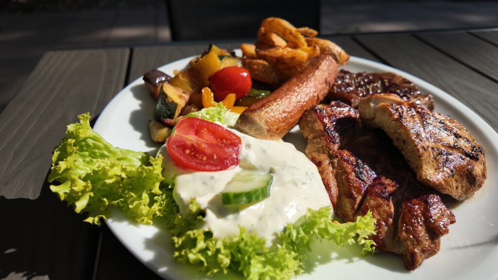On a white plate are grilled chicken, steak, French fries, fried vegetables, lettuce, a tomato slice, cucumbers and a creamy white sauce. Enjoy this lively outdoor meal - perfect for your next summer prepaid meal.