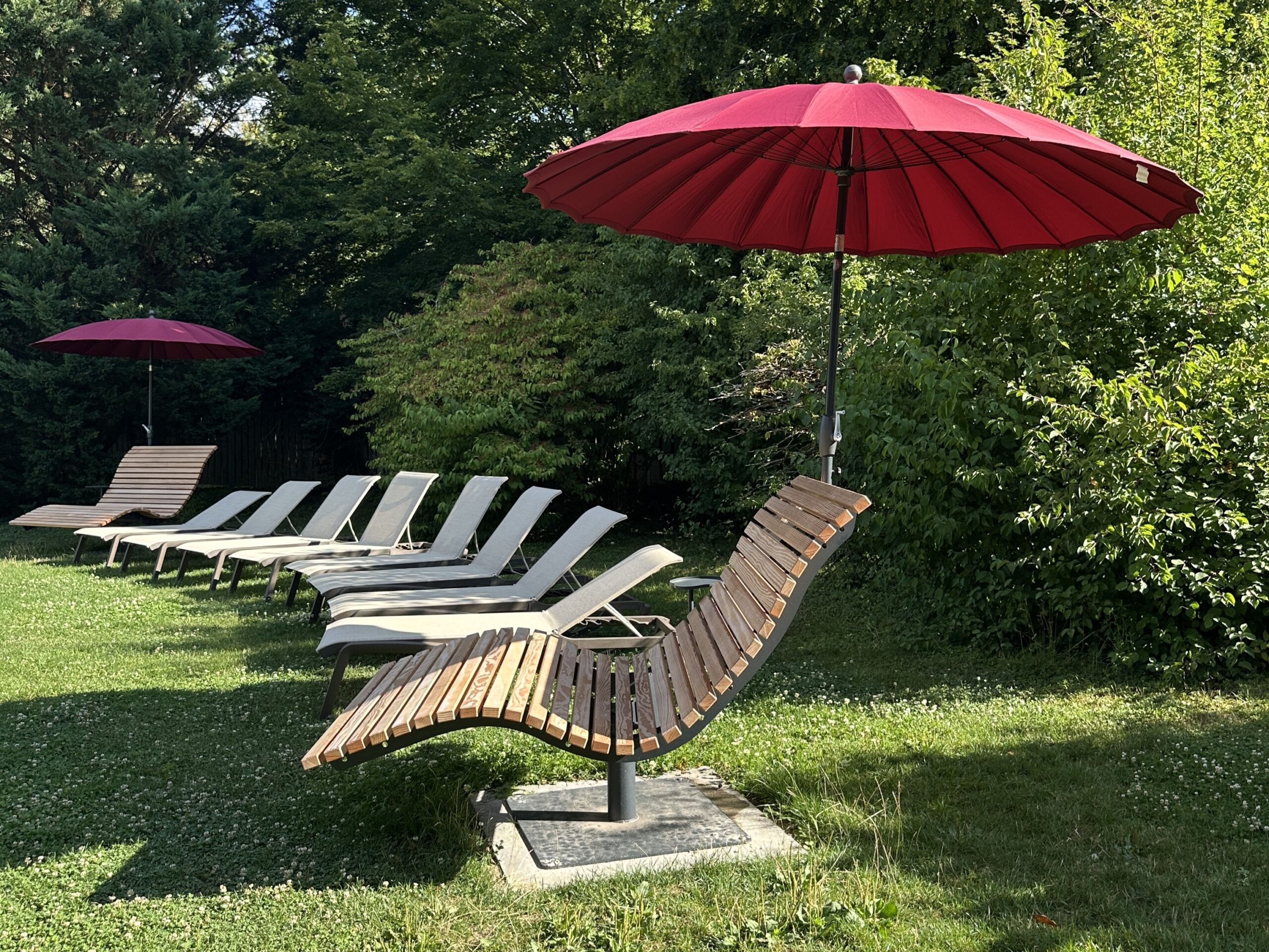 Several wooden lounge chairs with red umbrellas stand in a row on the grassy lawn, with lush green bushes and trees in the background. The inviting surroundings invite you to relax outdoors - an ideal place to enjoy your summer value card.