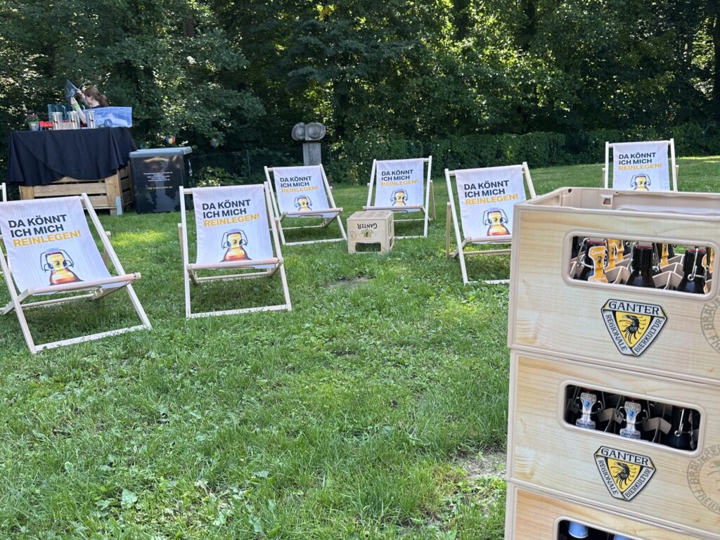 Several wooden lounge chairs with funny signs stand in the grass next to stacked Ganter beer crates and a summer prepaid card, in the background there are trees and a small drinks station - perfect for an outdoor event.