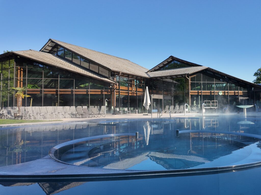 A modern building with large glass windows and wooden beams sits behind a steaming outdoor pool, where guests can relax on lounge chairs under blue skies -- perfect for relaxing with the summer prepaid card.
