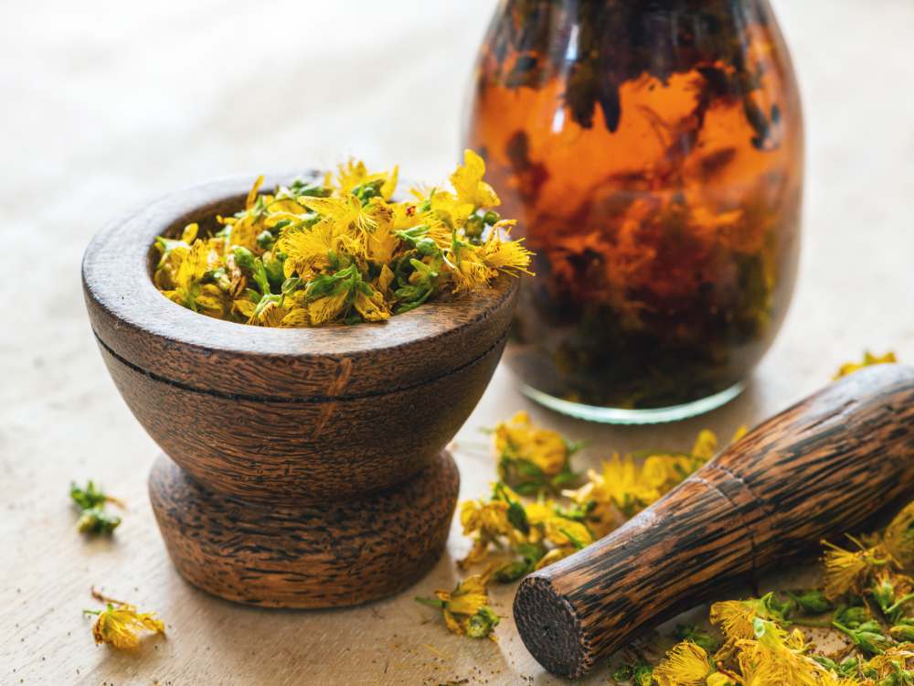 A wooden mortar filled with yellow dried flowers sits next to a matching pestle and a glass jar of amber liquid and floating herbs, all on a bright surface that exudes the natural charm of a summer value card.