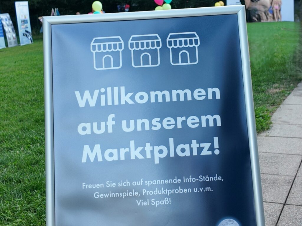 A sign on grass with icons of market stalls reads: Welcome to our marketplace! Look forward to exciting information stands, competitions, summer value cards, product samples and much more. Balloons can be seen in the background.