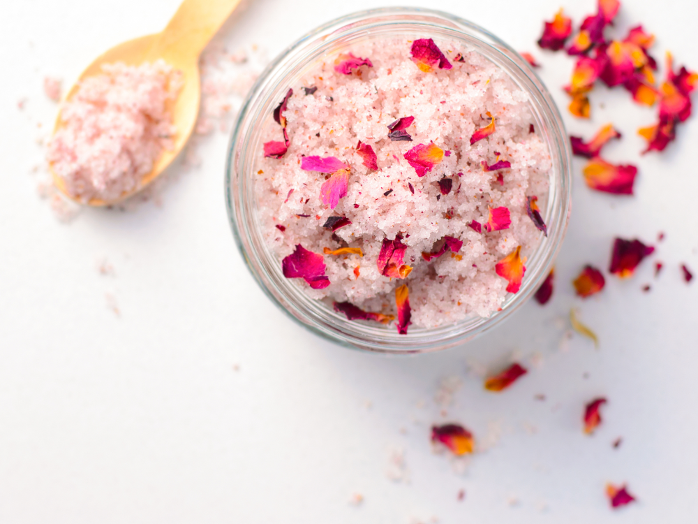 On a white surface stands a glass pot with pink bath salts mixed with dried rose petals, as well as a wooden spoon and scattered petals - the perfect self-care after enjoying the summer prepaid card.