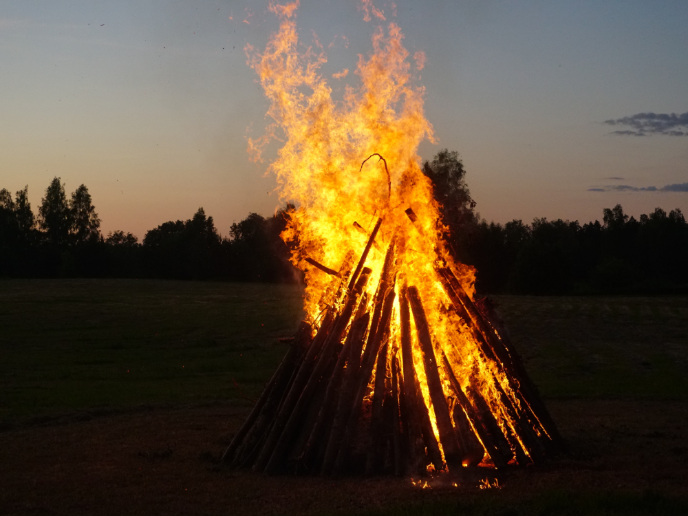A large campfire made of logs burns brightly on a meadow at dusk, the trees stand out against the evening sky - the perfect backdrop for your summer value card.