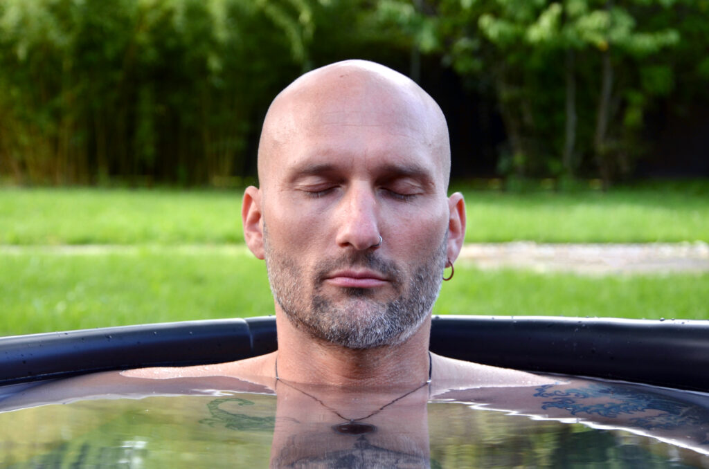 A bald man with a beard and a small earring sits with his eyes closed in an outdoor pool of water and relaxes. He embraces ice bathing in the Keidel Therme and finds peace while green grass and trees gently blur in the background.